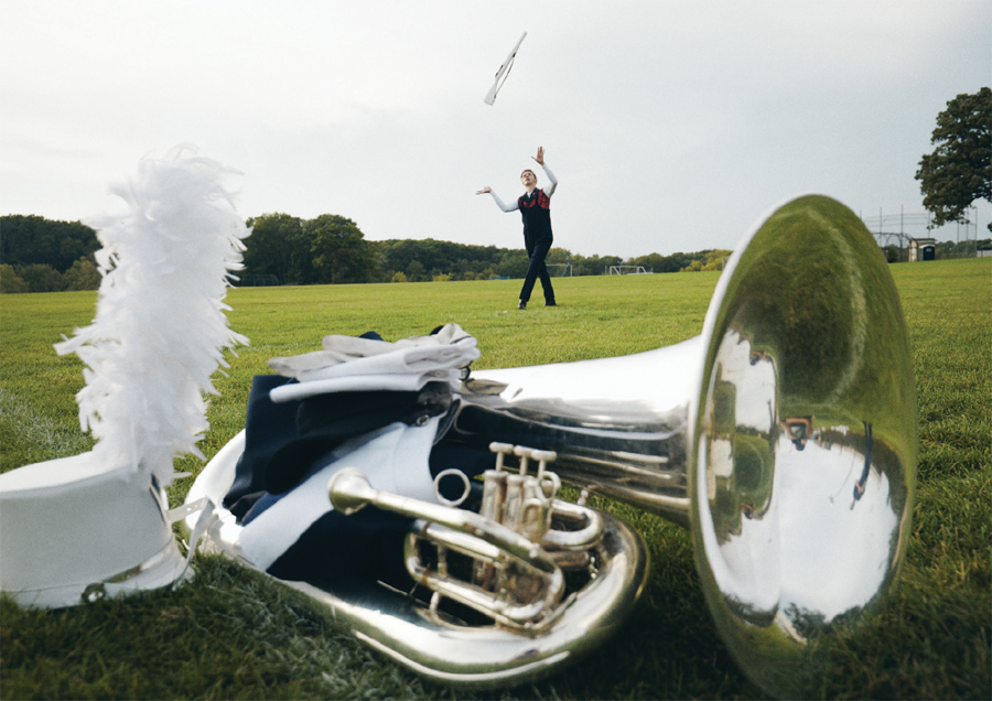 A tuba and marching band hat with plume rest in the foreground on a football field while a color guard member practices throwing a rifle in the background.