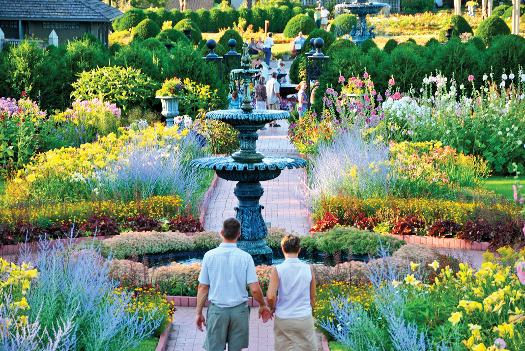 A couple walk through manicured flower beds and water features in the Munsinger Clemens Gardens.