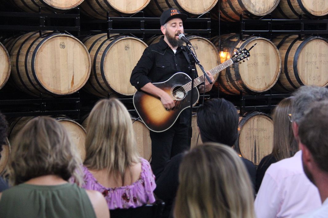 Ross Golan performs The Wrong Man album at Tattersall Distillery, a wall of barrels behind him and a few rows of audience members in front of him.