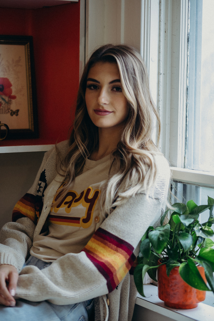 A brown-haired caucasian woman sitting near a window wearing a tee and sweater made by Hippy Feet